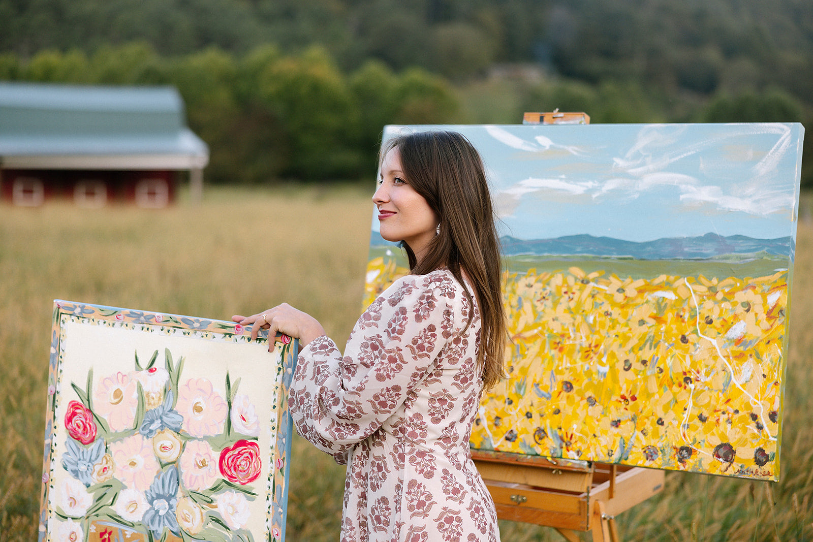 Woman holding two paintings of floral and sunflower scenes in a field.