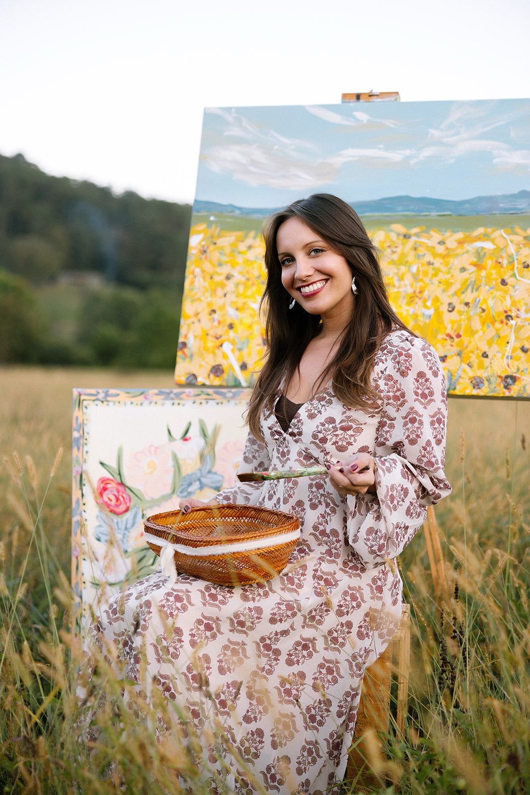 Woman in a floral dress holding a painting of a field with a large canvas behind her.