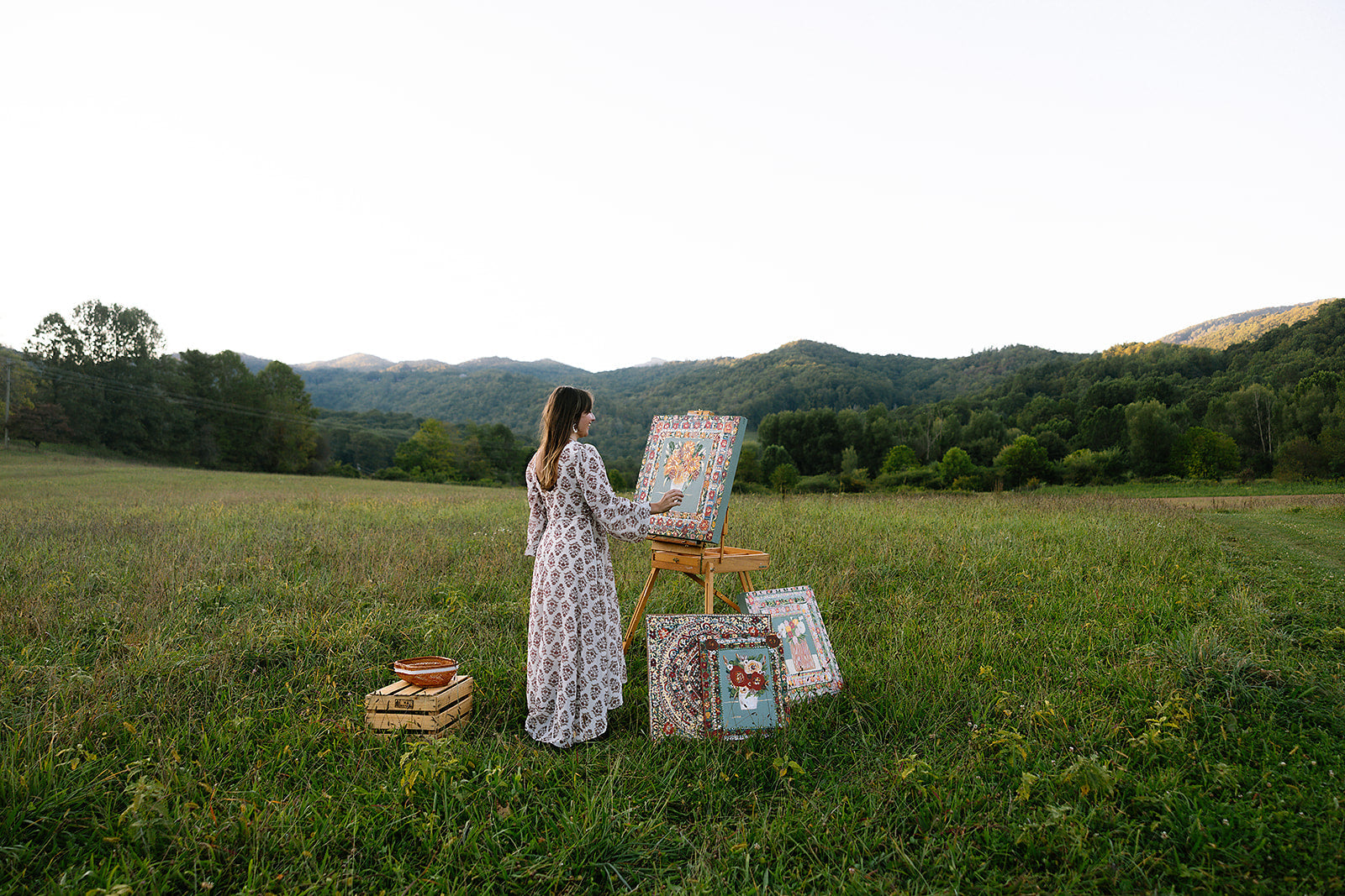 Woman painting outdoors in a scenic field with mountains in the background